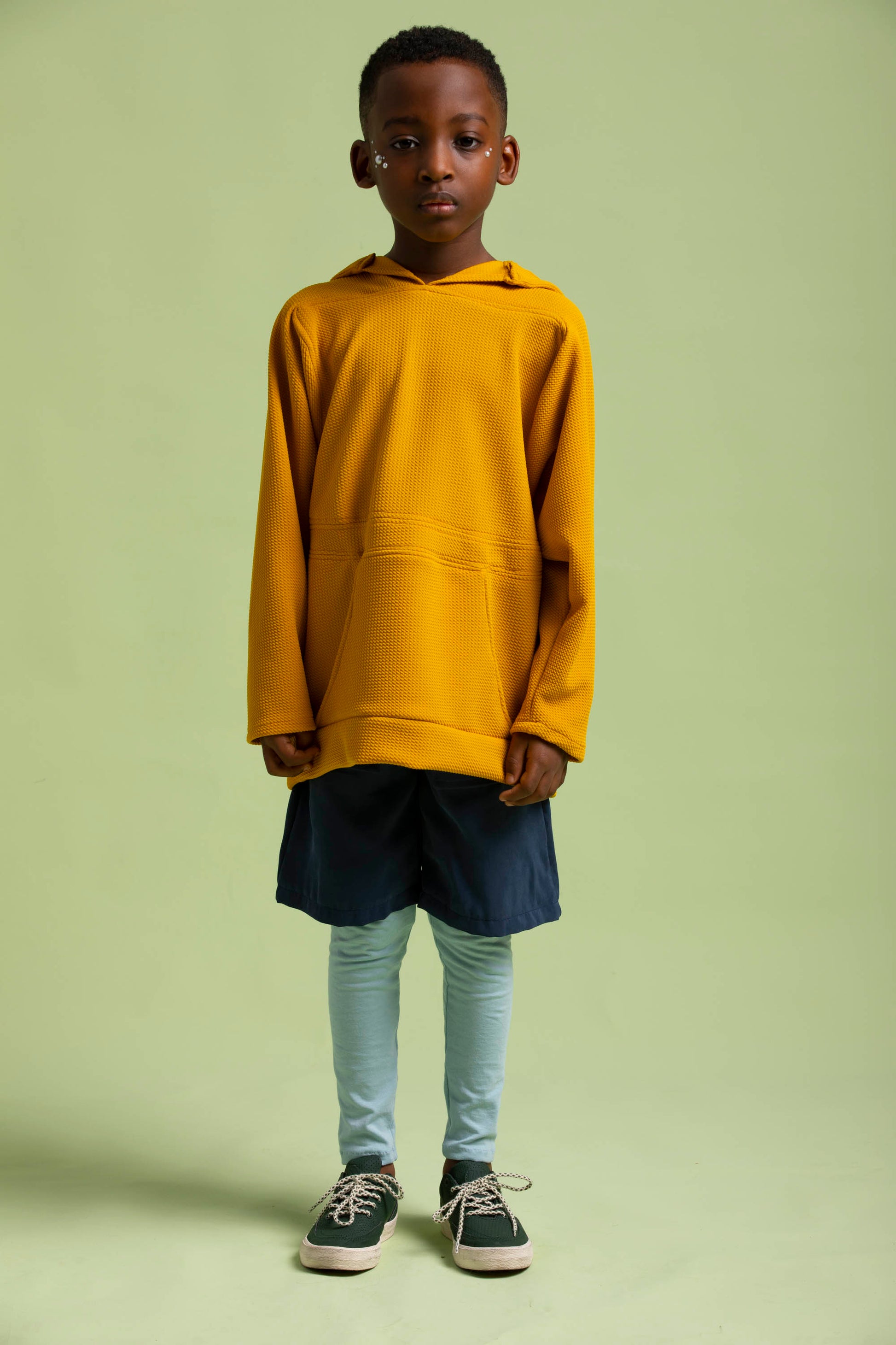 Young boy poses in a yellow embossed knit hoodie paired with navy fauno shorts layered over leggings