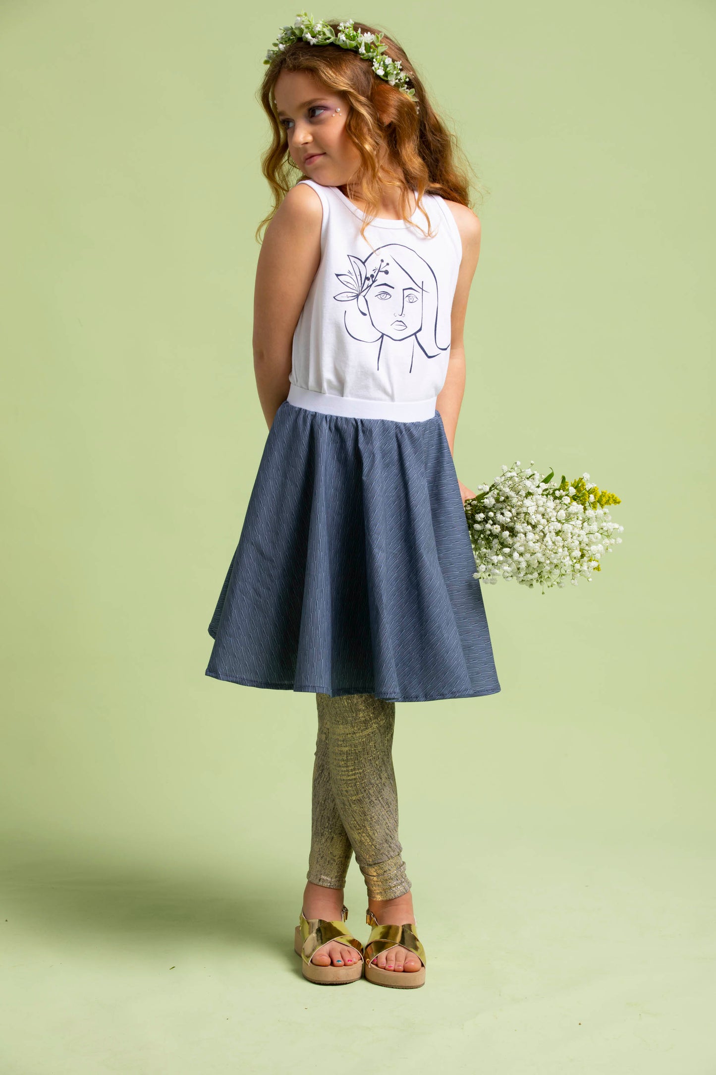 A young girl with flowers behind her back shows off her Ofelia T-Shirt with a navy Mercedes Skirt